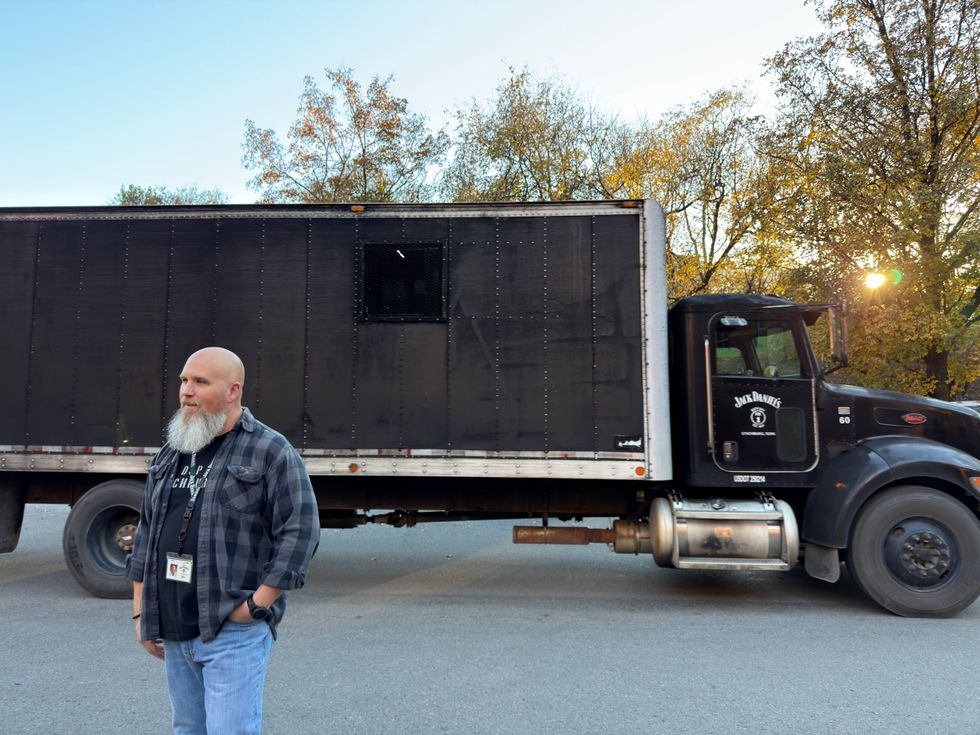A bearded man stands in front of a large black truck with "Jack Daniels" branding. Trees with autumn leaves are in the background, with a setting sun.