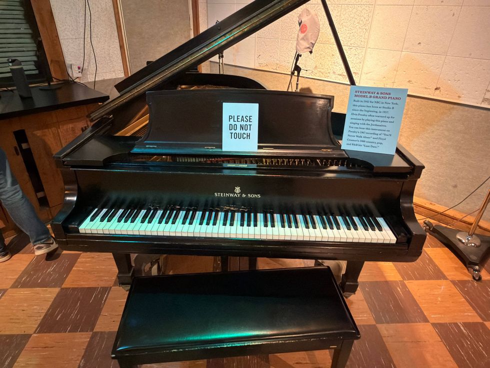 A black Steinway & Sons grand piano sits in a studio with a "Please Do Not Touch" sign on it. The room has wooden floors and acoustic panels.