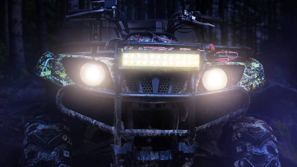 a camouflaged dirty offroad hunting atv vehicle standing at night in forest with headlights turned on, front view.