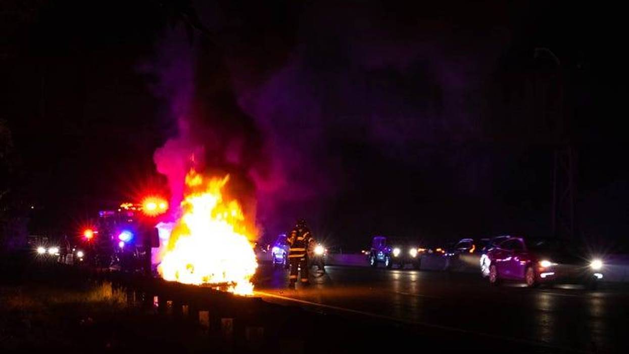 A car on fire on a highway at night. Police are behind the wreck. Firefighters stand to the side, working to douse the fire. Other cars pass by on the side.
