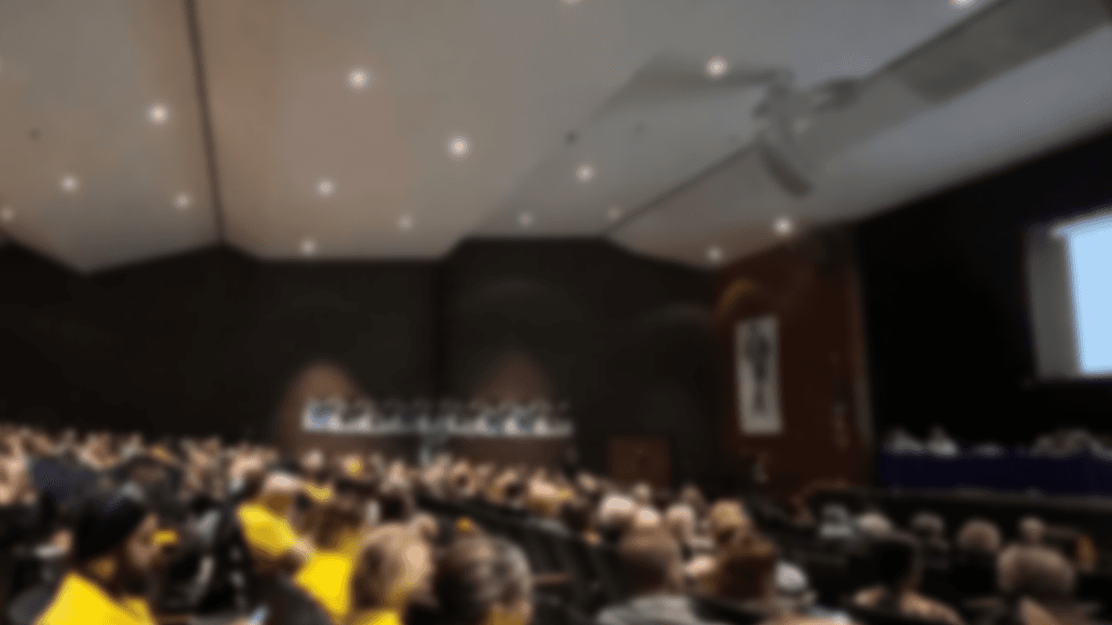 A crowd sits in an auditorium. Some people are wearing yellow. They are focused on a presentation on the stage on the right side of the picture.