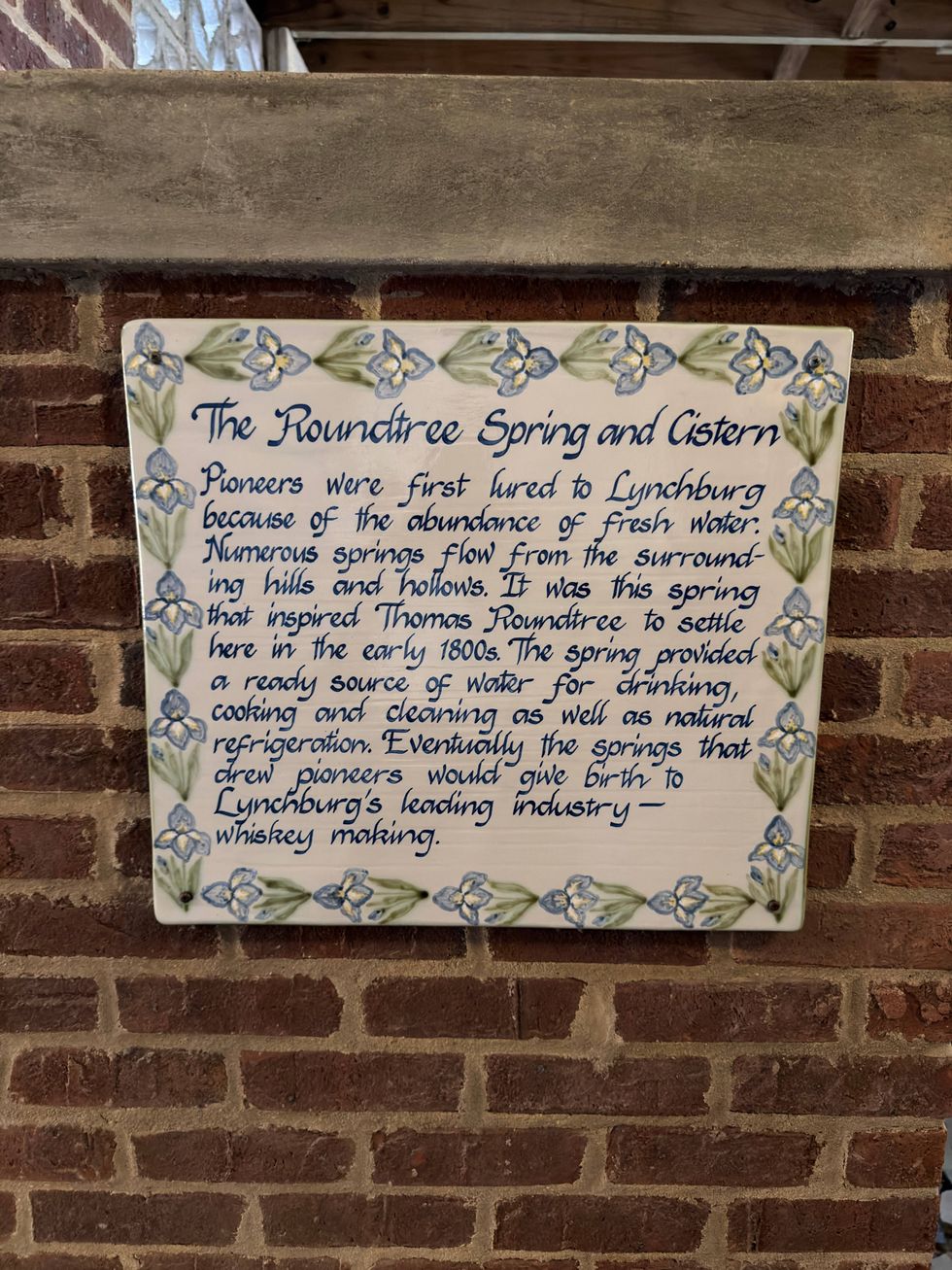 A decorative historical plaque on a brick wall details the significance of Roundtree Spring in Lynchburg. Blue and green floral patterns adorn the border.