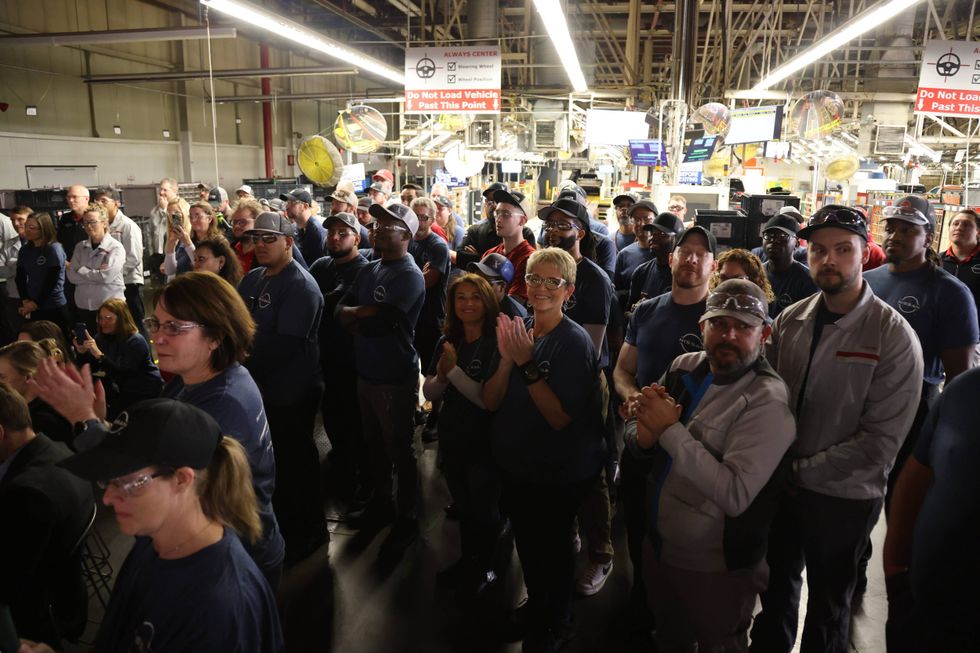 A diverse group of factory workers stands together in a brightly lit industrial space, some clapping. The mood is attentive and engaged. Signs and machinery are visible in the background.