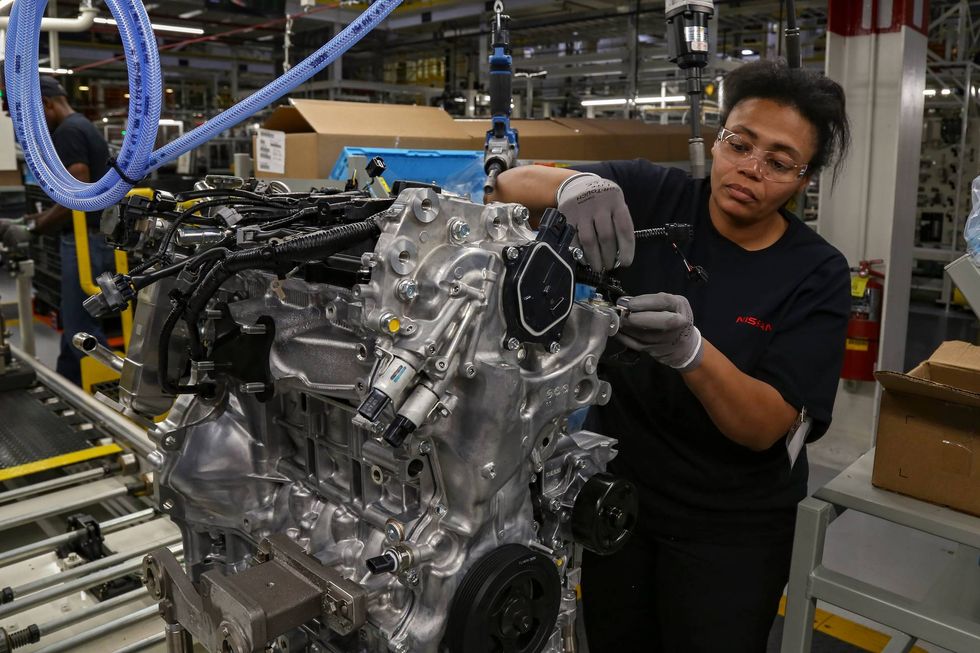 A factory worker meticulously assembles a silver engine on an industrial floor. She wears goggles and gloves, conveying focus and precision in her task.