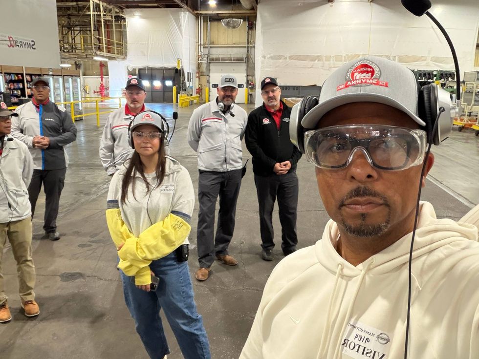 A group of people in safety gear, including hard hats and goggles, stand inside an industrial warehouse. The mood appears focused and professional.