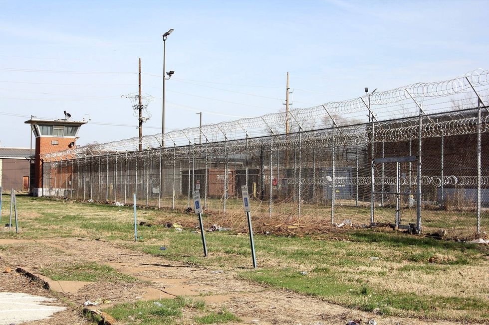 A guard tower and courtyard at a jail. Wire fences surround brick buildings in the background.