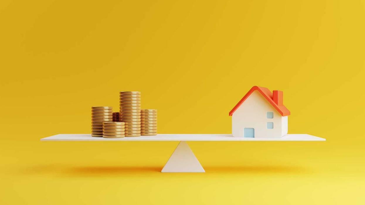 A little house and coins sitting on a balance beam.