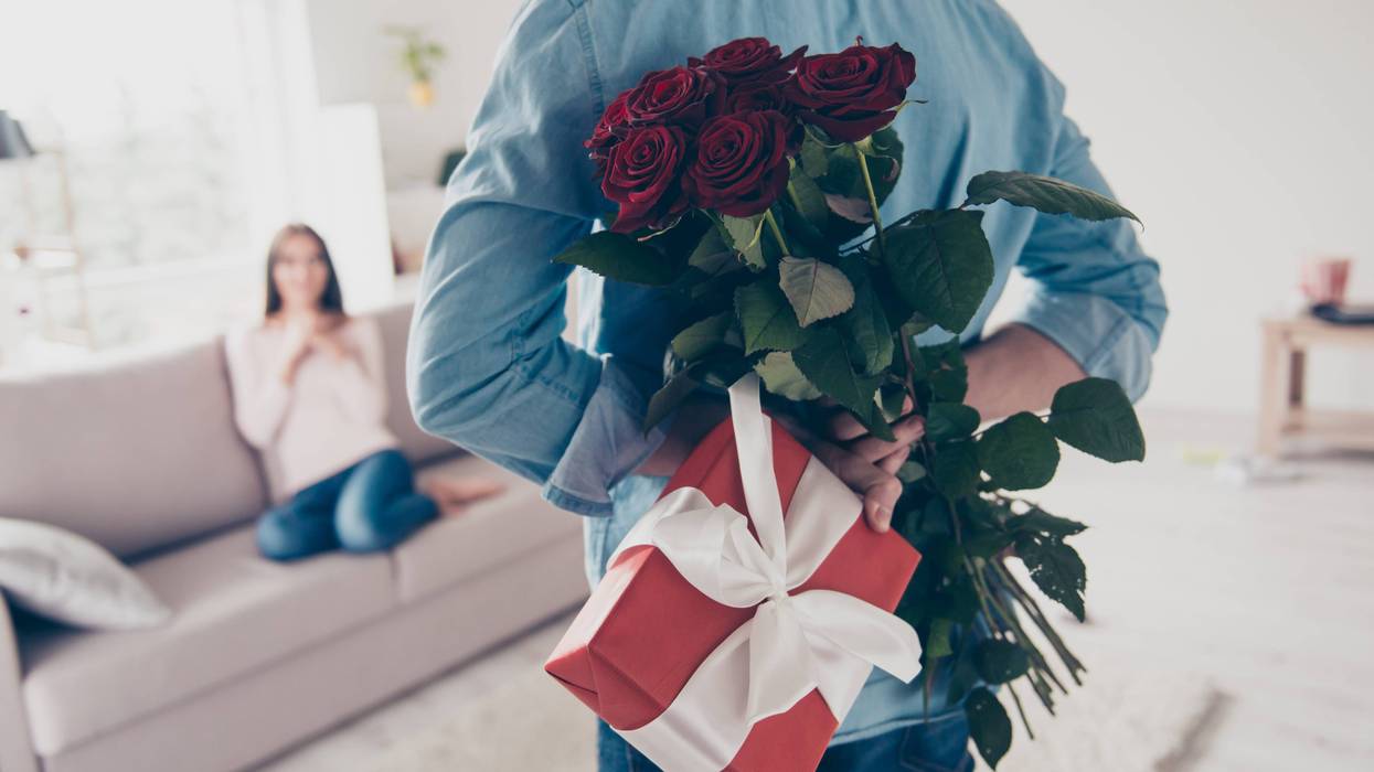 A man holds a wrapped gift and a bouquet of flowers behind his back while a woman waits on a couch in the background