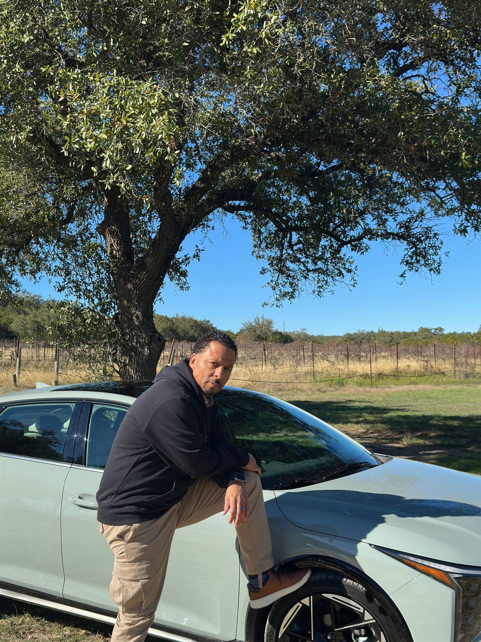 A man leans against a car in a sunny field, surrounded by green grass and open space.