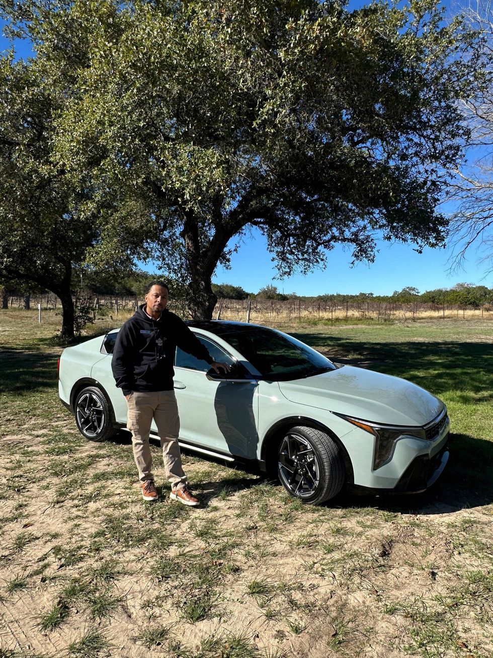A man stands beside a green car in a sunny field, surrounded by grass and open space.