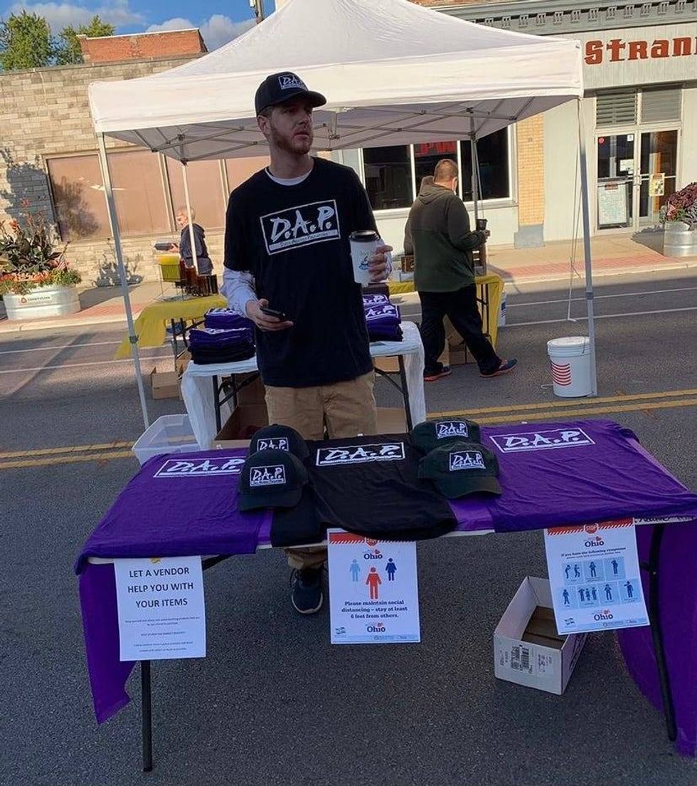 A man wearing a D.A.P t-shirt standing behind table of D.A.P. merchandise