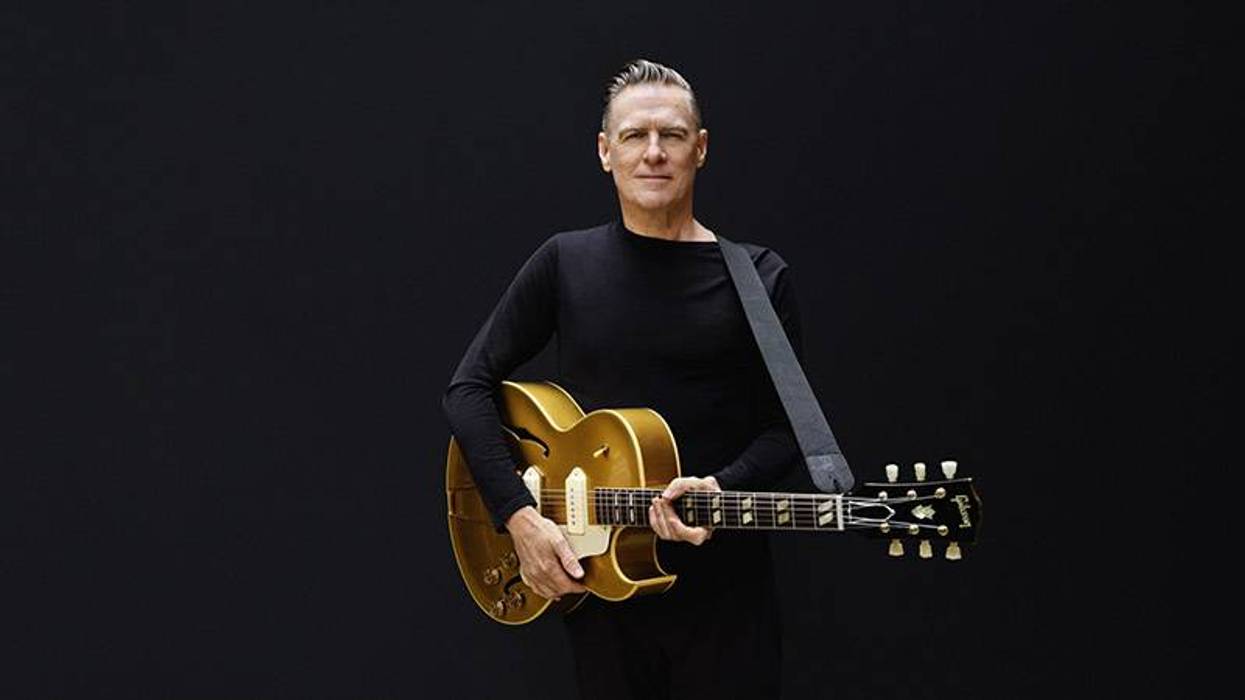A man with grey hair, wearing a black shirt, holds a gold electric guitar against a dark background.