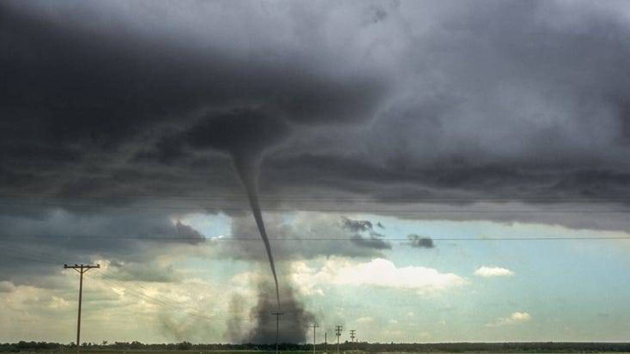 A powerful tornado descends from dark, stormy clouds, kicking up dust in a flat rural field.