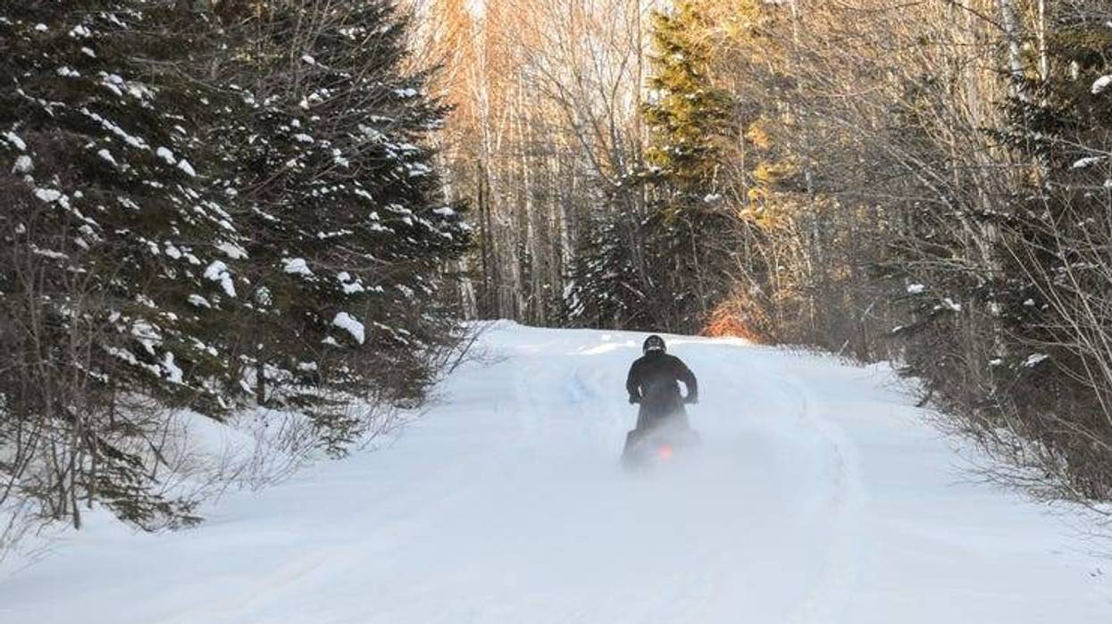 A snowmobiler seen driving away through a clearing in a wooded area.
