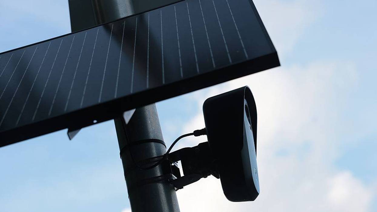 A solar panel and black camera mounted on a pole against a blue, cloudy sky.