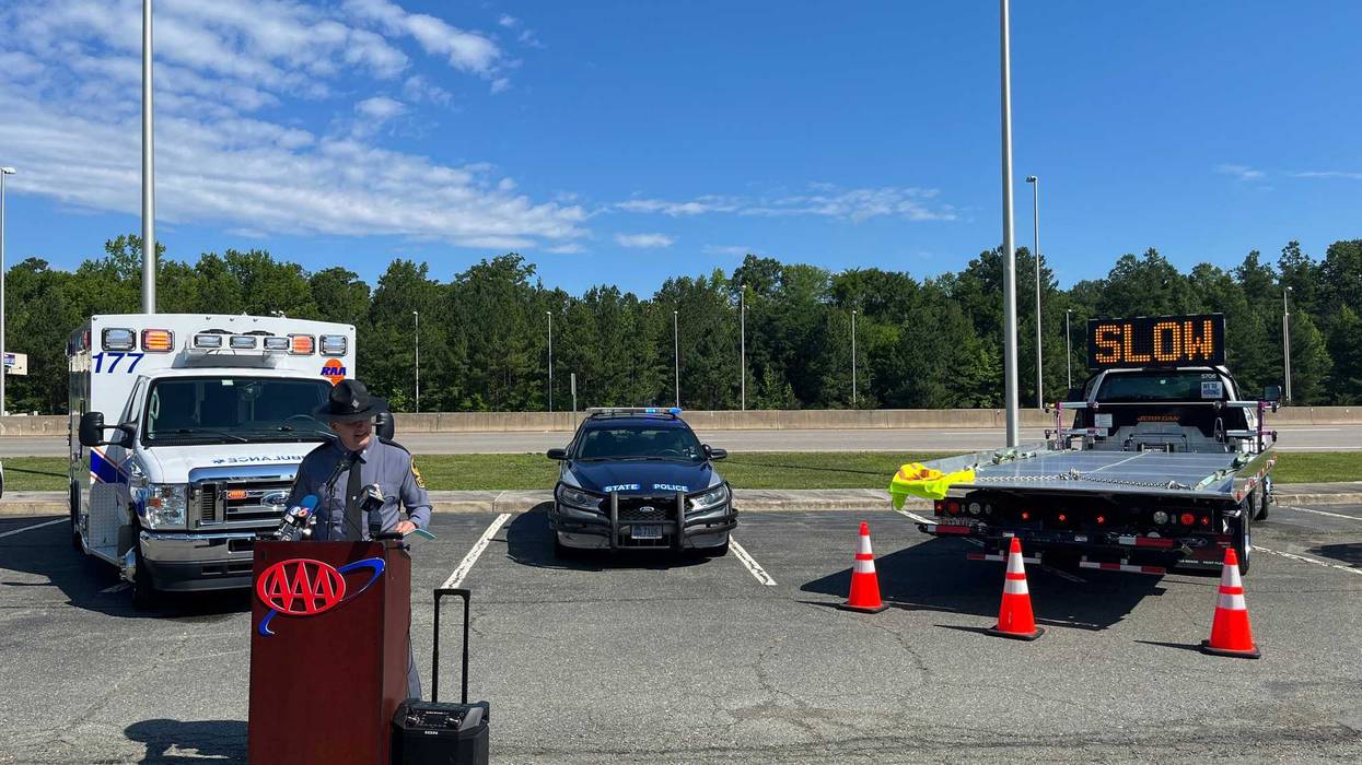 A State Trooper speaks at a lectern in front of an ambulance, a police car, and a flatbed.