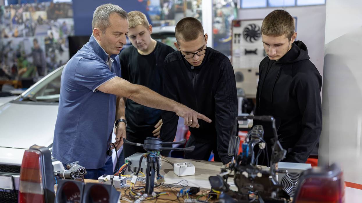 A teacher mentors a trio of male students learning automotive electronics.