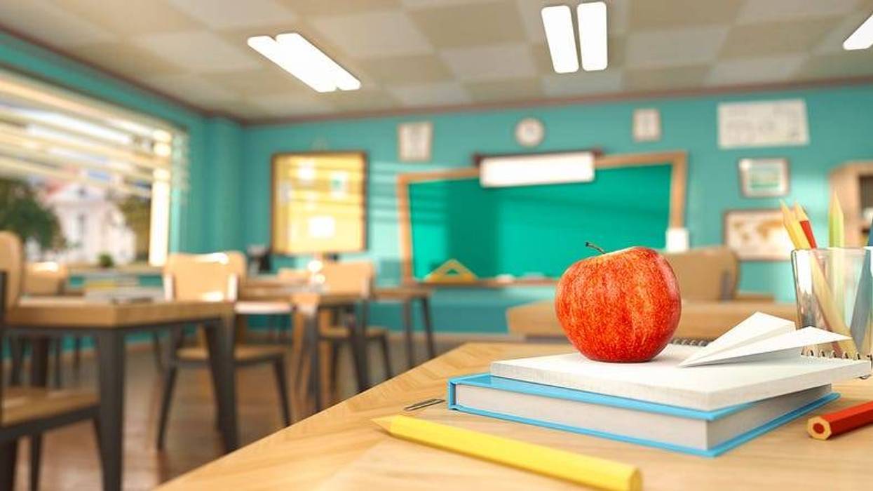 A teacher's desk with a textbook, pencils, paper and an apple on top. The rest of the classroom, student desks, a chalkboard and a bulletin board are out of focus in the background.