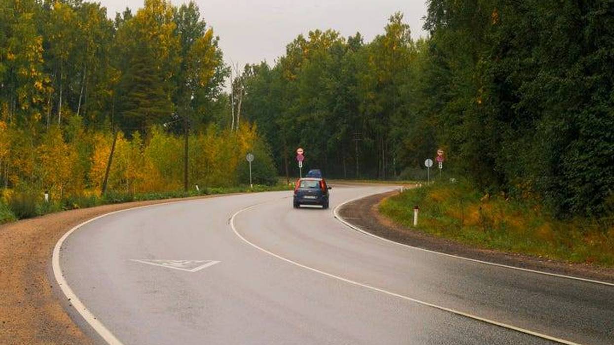 A two-lane rural highway with forested area on both sides. A couple of cars can be seen going around a curve.