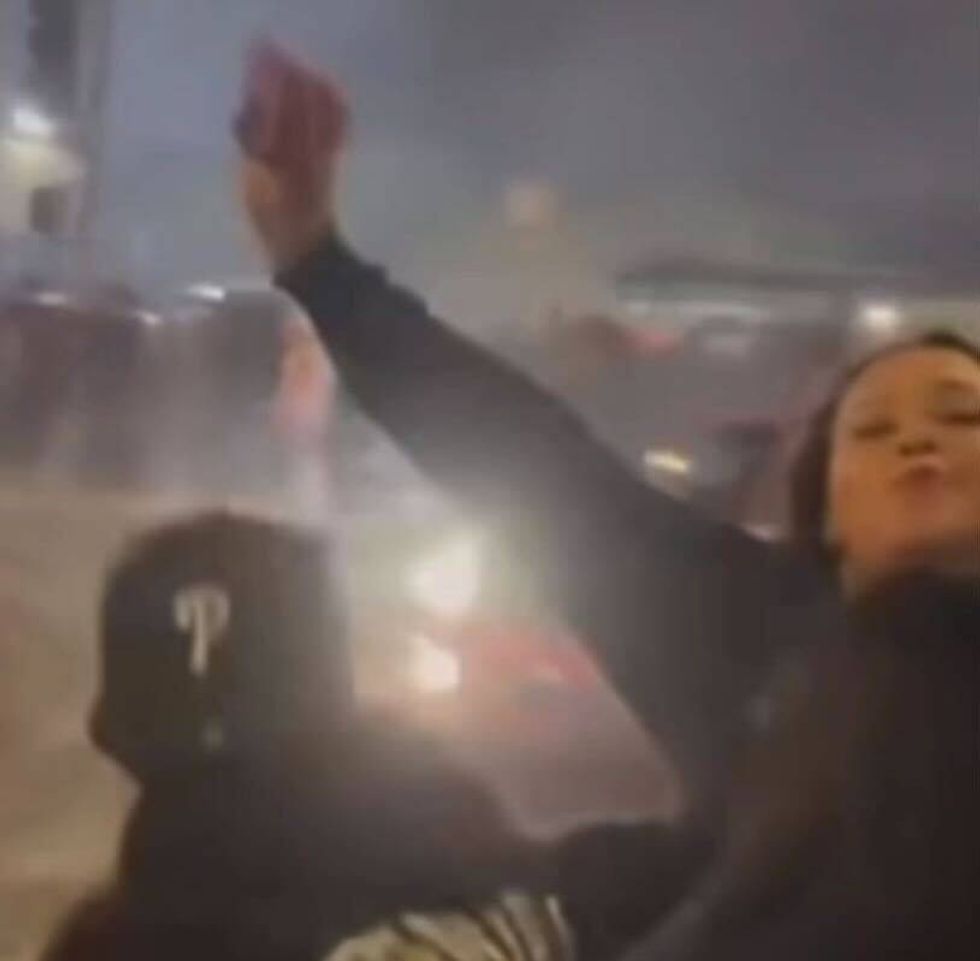 A woman in a dark sweatshirt raises her hands in the air as a car drives in the background