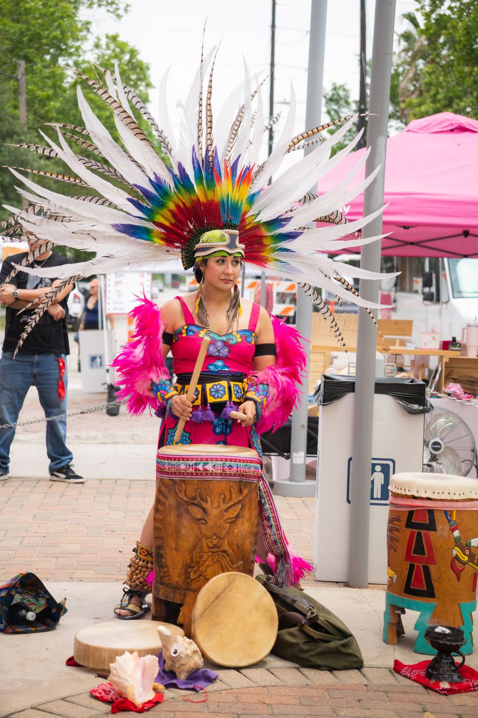 A woman in a huge white and rainbow feathered headdress, pink dress, playing a carved drum outdoors.