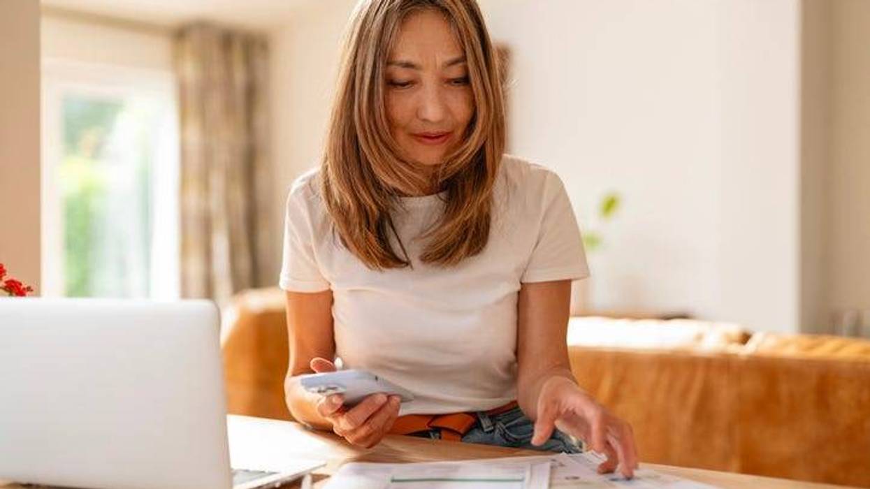 A woman is sitting at a wooden table, holding a smartphone and sorting through papers in a well-lit home office filled with natural light.