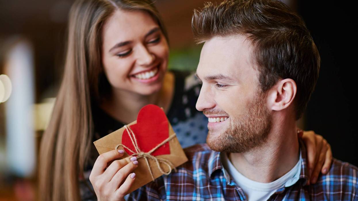 A woman leans over a man's shoulder to hand him a gift wrapped in brown paper with a large red paper heart affixed to it