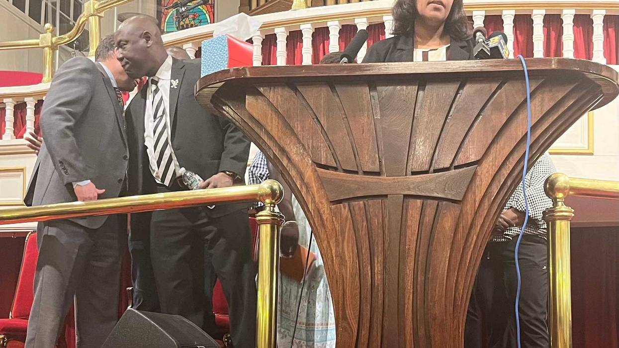 A woman stands at a lectern, speaking. Several men and women stand behind her.