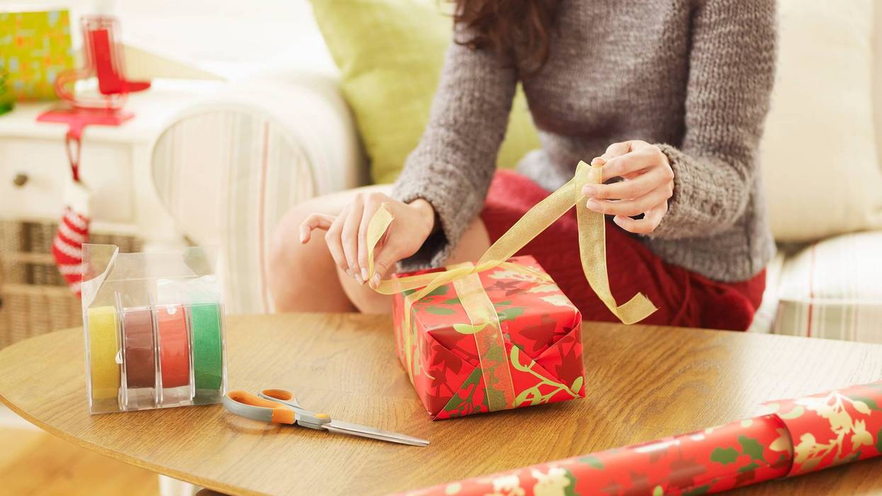 A woman ties a bow on a wrapped present with tape, scissors and a roll of wrapping paper lying on the table nearby