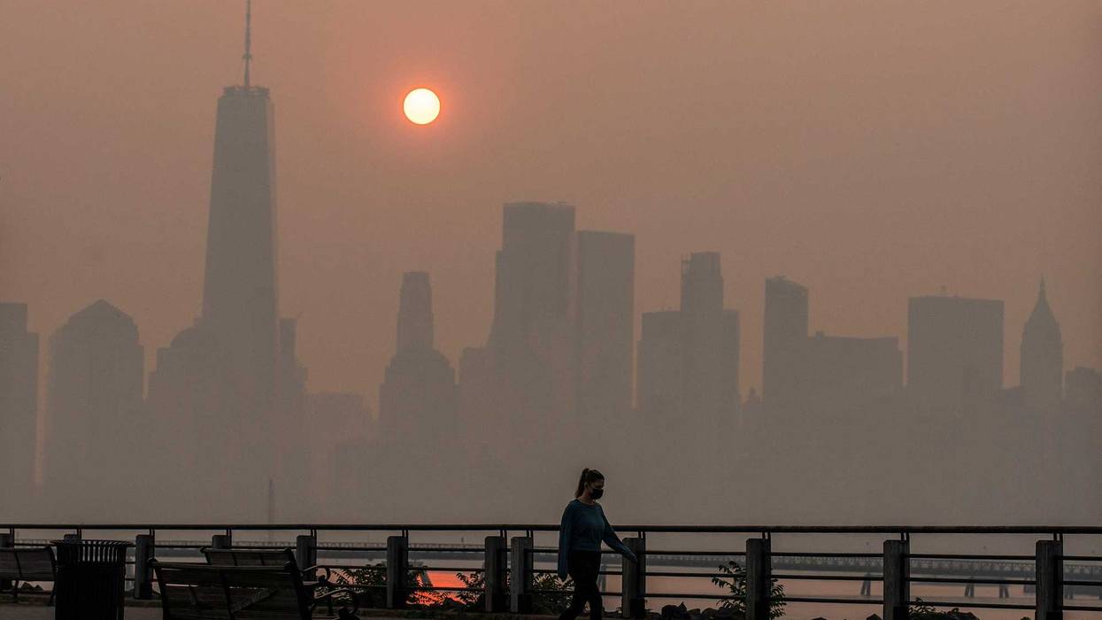 A woman walks as the sun rises behind the One World Trade Center and the New York skyline, while the smoke from Canada wildfires covers the Manhattan borough as it is seen from the Liberty State Park on June 8, 2023 in New Jersey