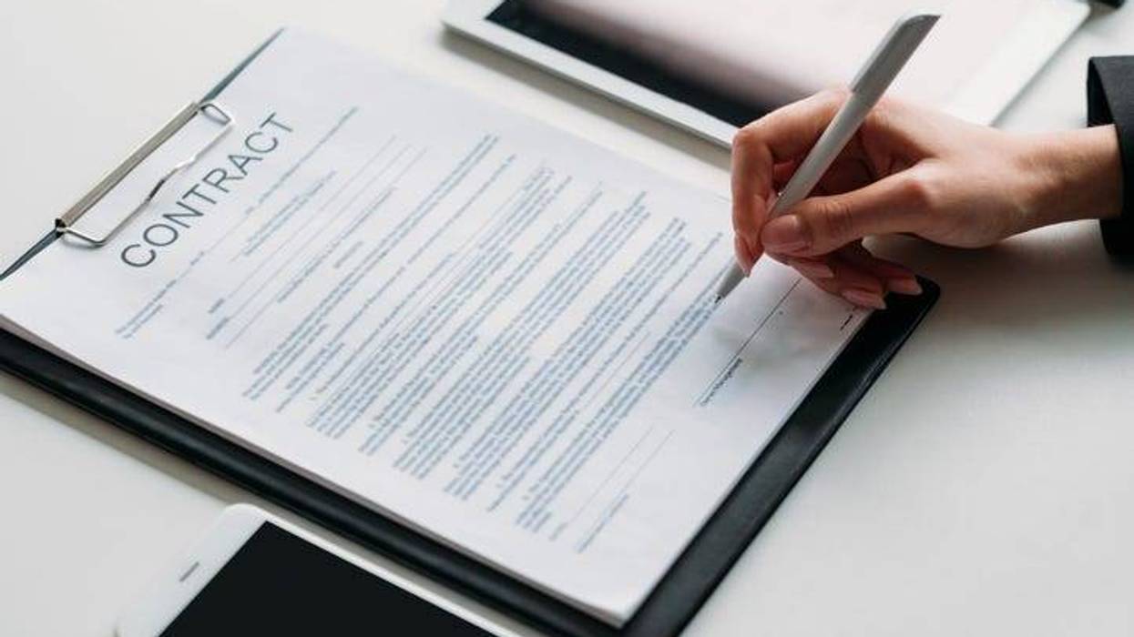 A written contract on a clipboard. A hand holds a pen and is poised to sign the contract.