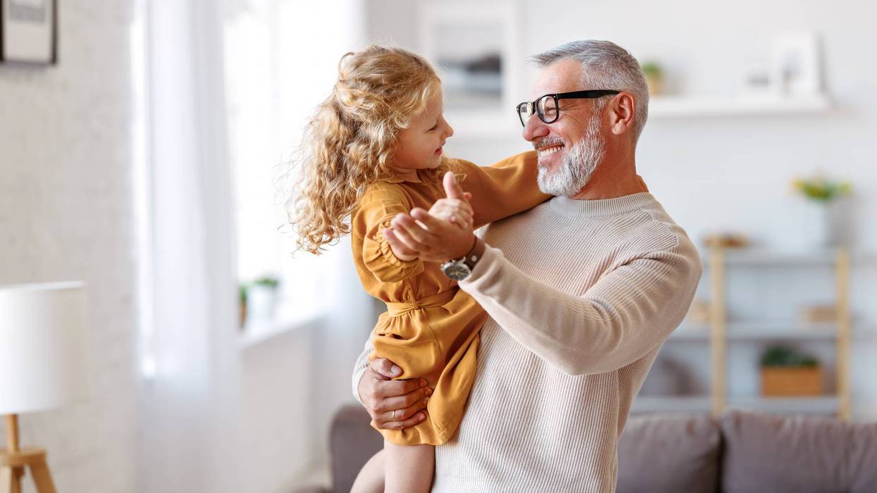 Adorable child girl and positive grandpa holding hands while dancing together in living room