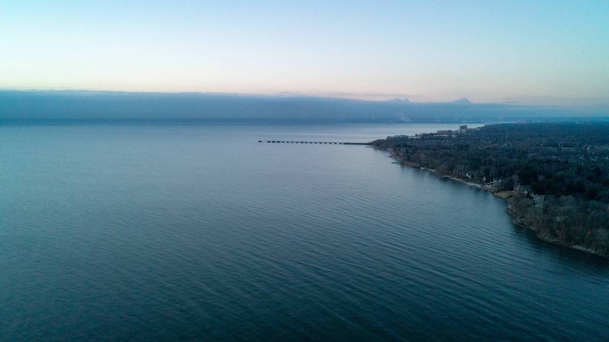 Aerial view of a calm blue lake with a long pier, a forested coastline, and distant hazy land.