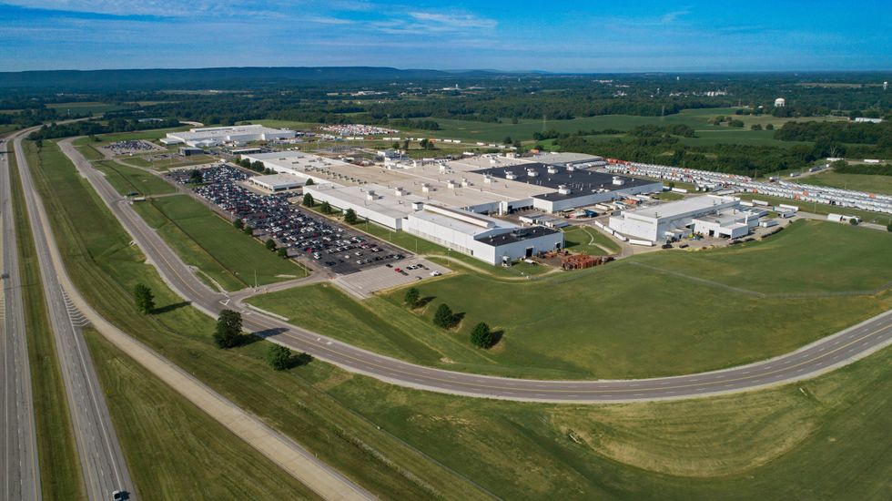 Aerial view of a large industrial complex with multiple white-roofed buildings and parking lots, surrounded by green fields and roads under a clear blue sky.