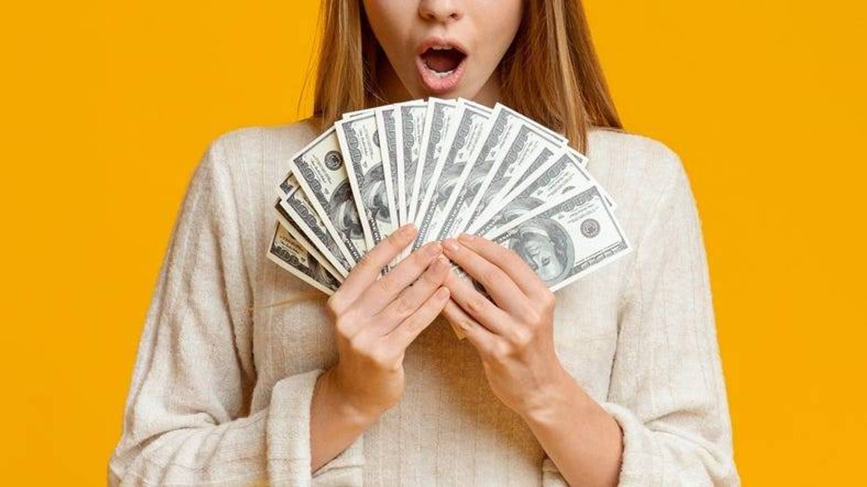 Amazed young woman holding bunch of money banknotes, celebrating profit over orange background, panorama with copy space
