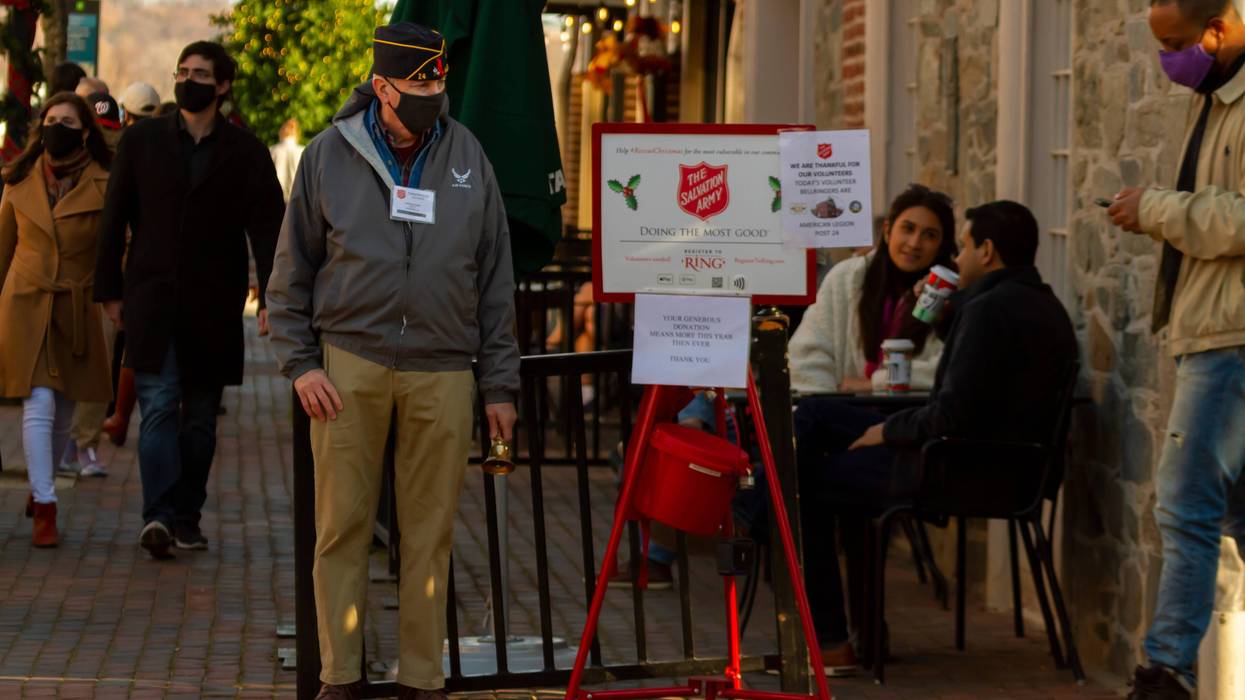 An elderly veteran with an airman side cap hat and face mask is standing next to a Salvation Army donation bucket, as a volunteer bell ringer.