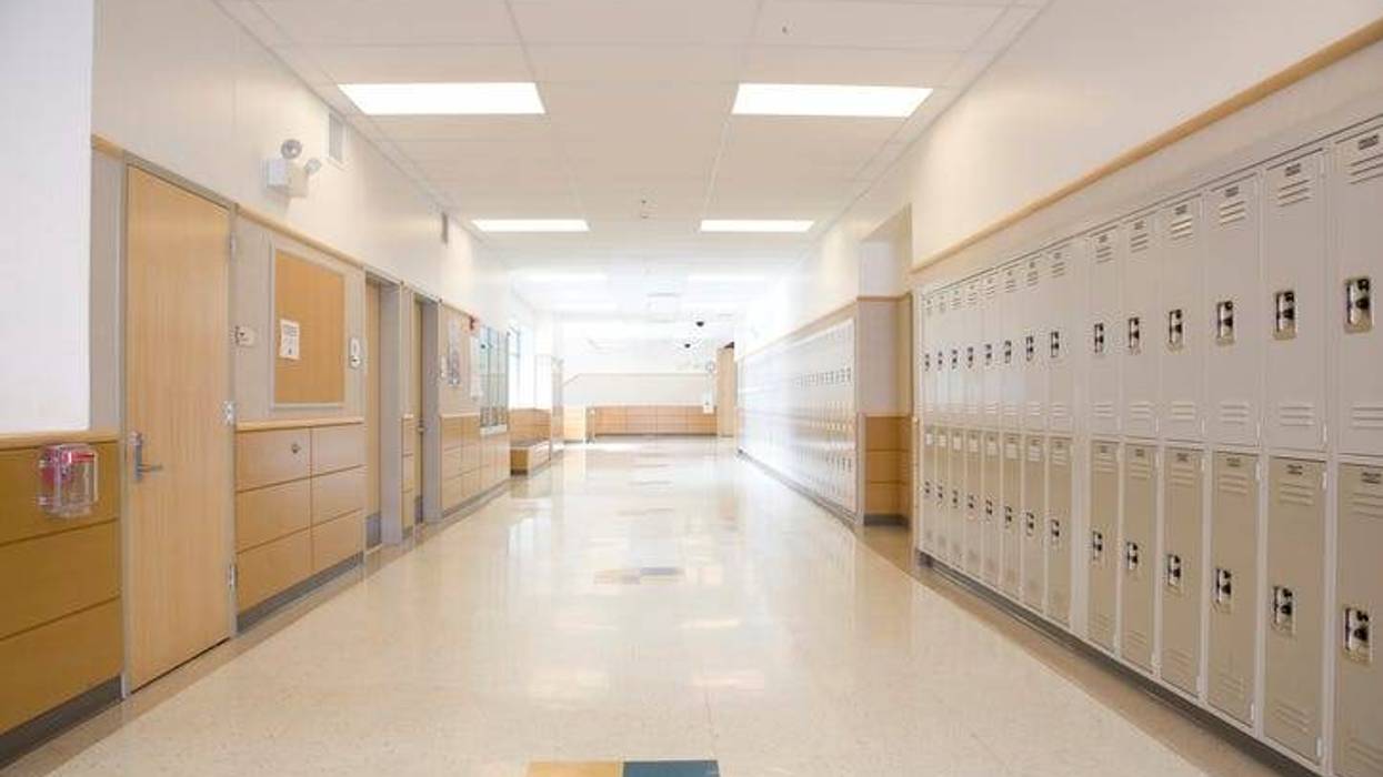 An empty school hallway lit by fluorescents. Lockers line one wall. Classroom doors and bulletin boards line the other wall.