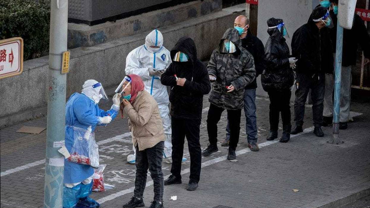 An epidemic control worker wears PPE to prevent the spread of COVID-19 as he gives a nucleic acid test to a resident as others wait in line wearing face shields before leaving by bus from an area with communities in lockdown on November 30, 2022 in Beijing, China.