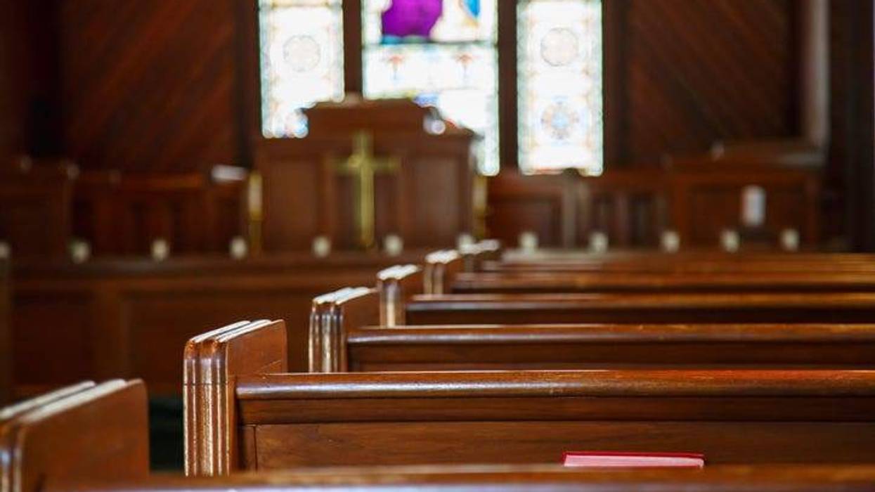 Angled shot of a church interior. Rows of pews face an altar. There's a stained glass window at the front.