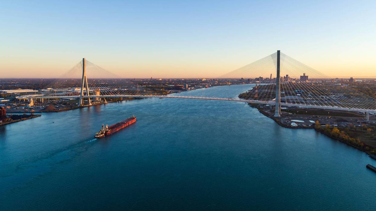 Barge on river under a large cable-stayed bridge with city skylines at golden hour.