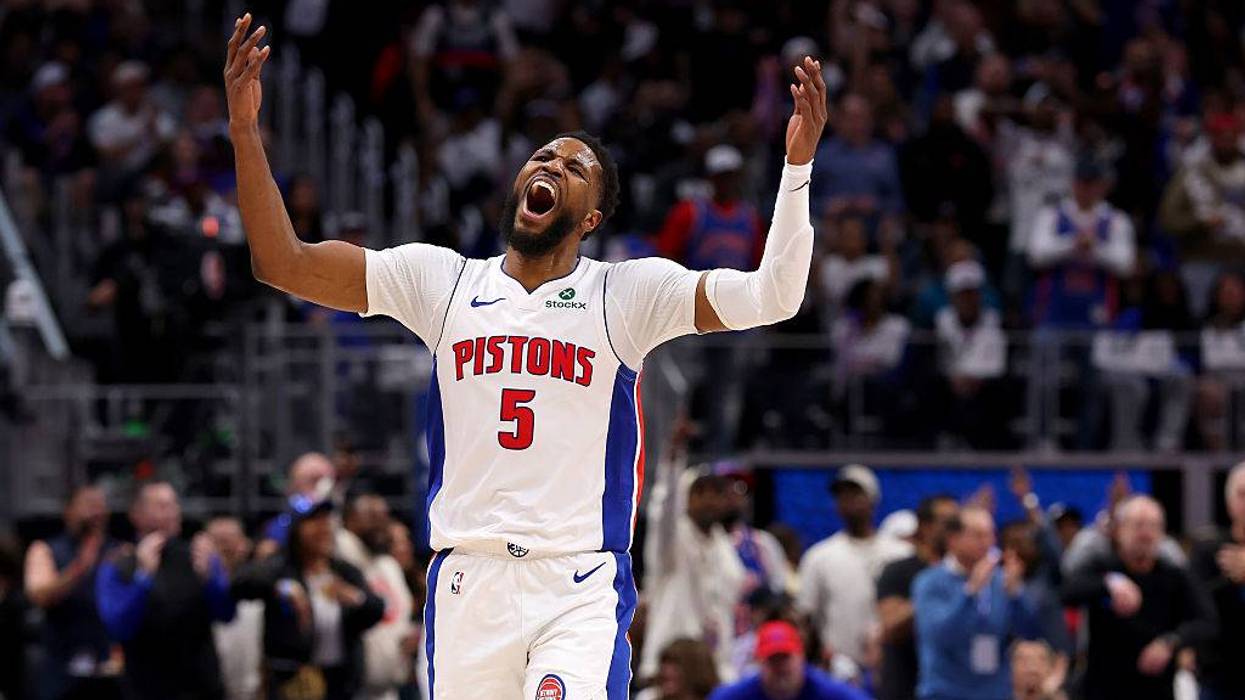 Basketball player in white Pistons #5 jersey raises arms and shouts intensely on court.