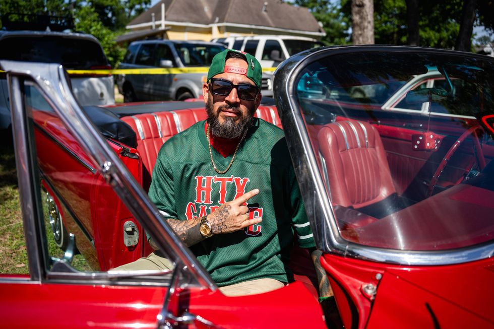Bearded man in sunglasses, cap, & tattoos in a red lowrider convertible, flashing peace sign.