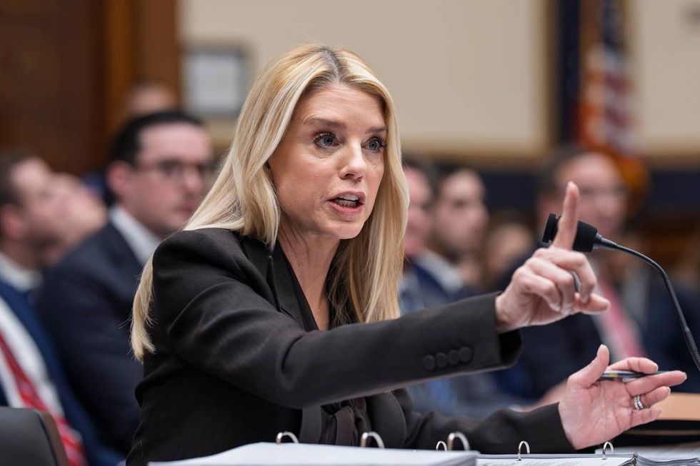 Blonde woman in black suit speaking, pointing finger near microphone at a hearing.