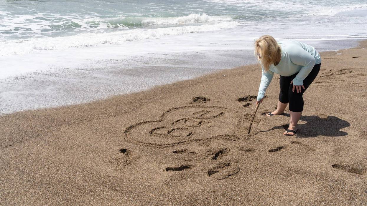 Blonde woman writes Bae (slang for Before Anyone Else, representing a romantic relationship) in sand on the beach