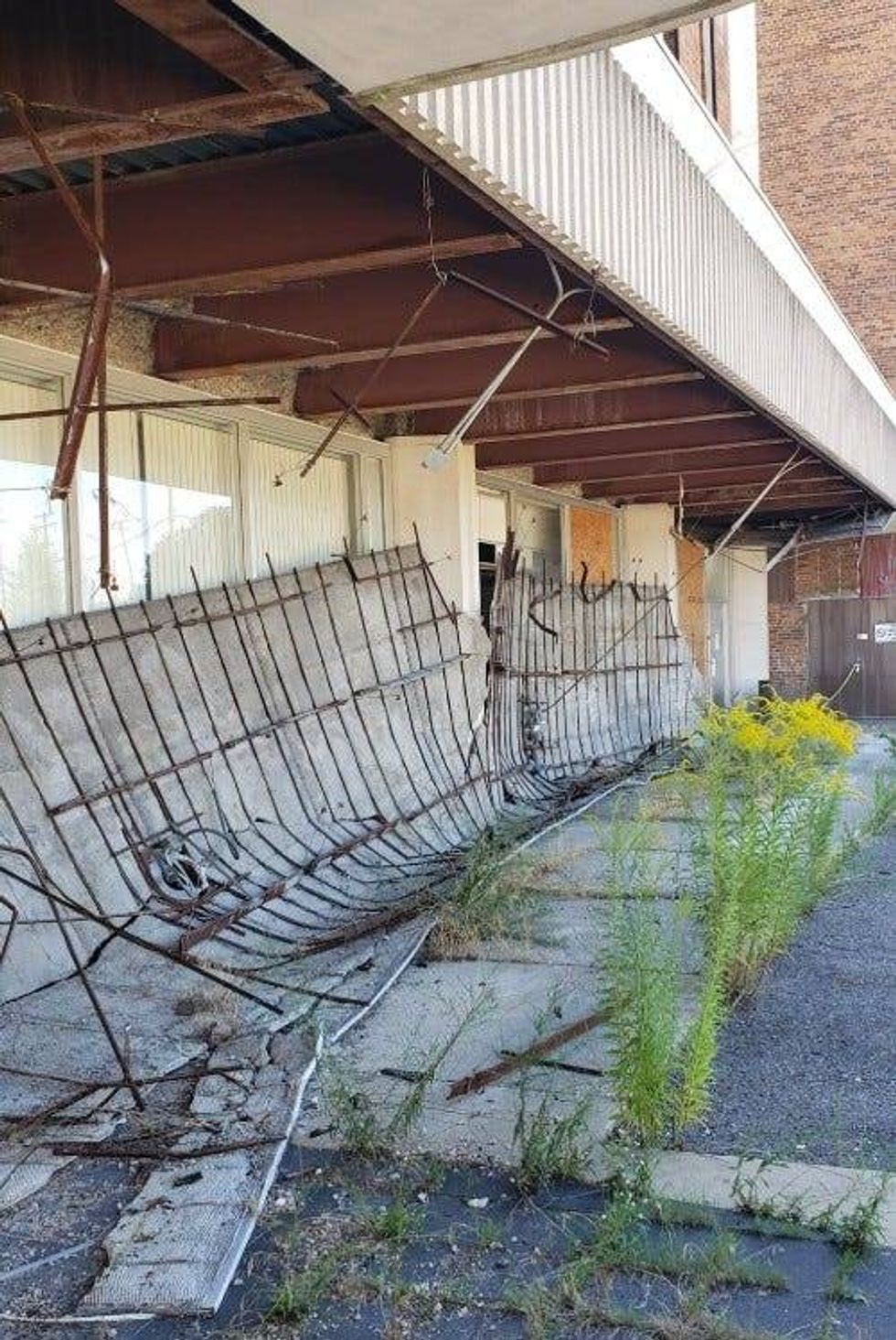Broken down fencing leans against the lower floor of the office building. The parking lot next to it is broken up, uneven and overgrown with weeds.