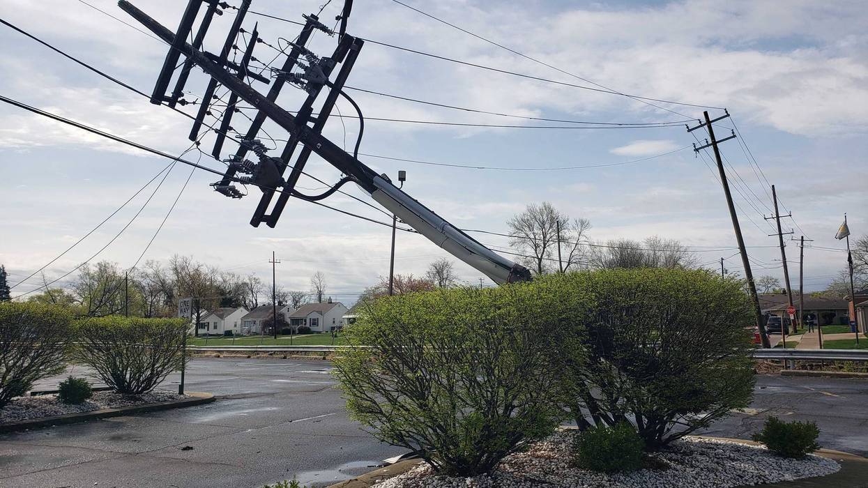 Broken utility pole leaning heavily, power lines sagging over a street. Houses in background.