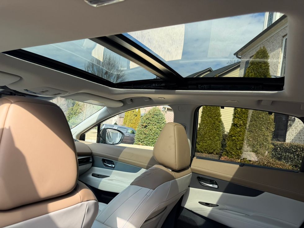 Car interior with panoramic sunroof, tan and white seats, looking out at trees and houses.