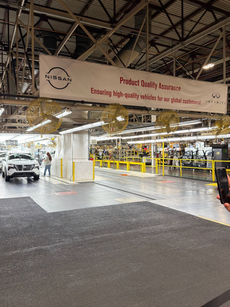 Car manufacturing plant with a Nissan banner reading "Product Quality Assurance." A worker stands near a white car. Industrial setting, efficient atmosphere.