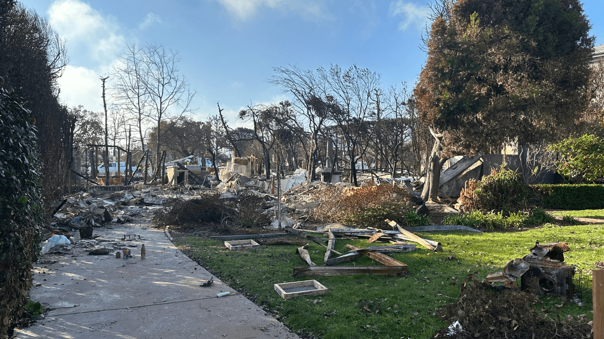 Charred debris and remnants of a destroyed home litter a sidewalk and lawn following the 2025 wildfire in Pacific Palisades, California. Burned vegetation, blackened trees, and fragments of framing and household items mark the path through what was once a residential area.