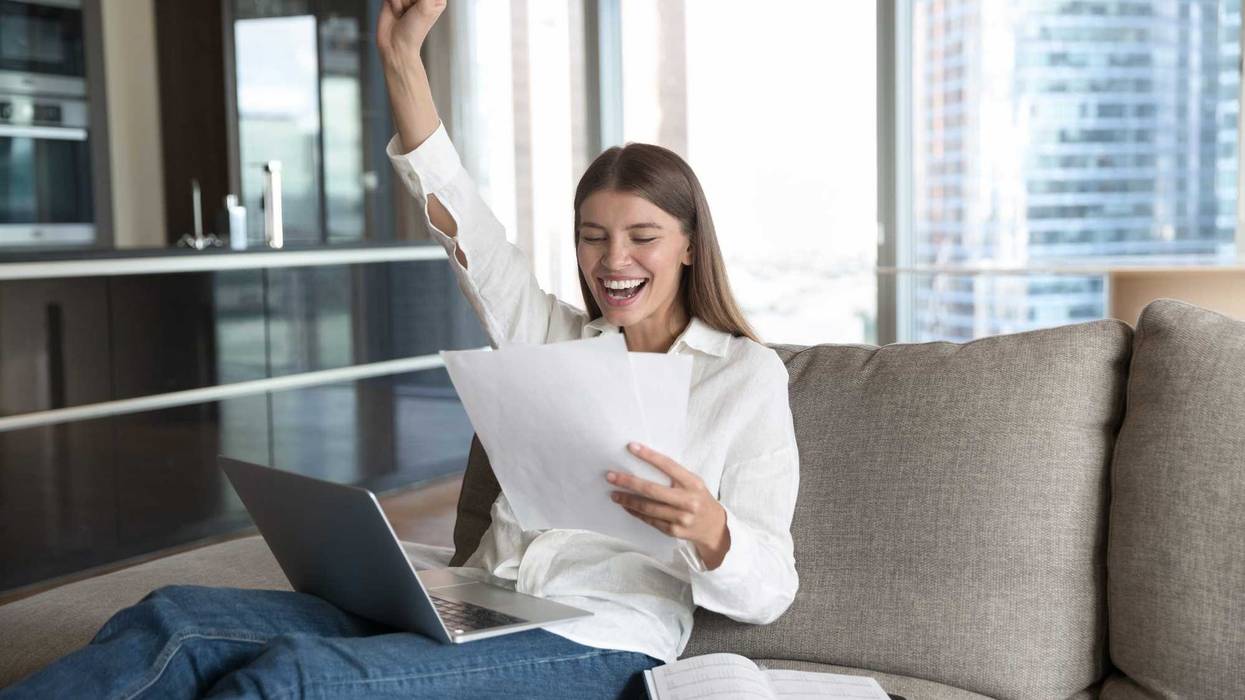 Cheerful young student woman excited with good news, reading paper document at laptop, resting on sofa at home, making winner hand, fist, smiling, celebrating success, laughing